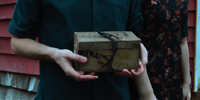 A picture of John and Lydia standing in front of an old, abandoned cabin with a mysterious box in their hands.